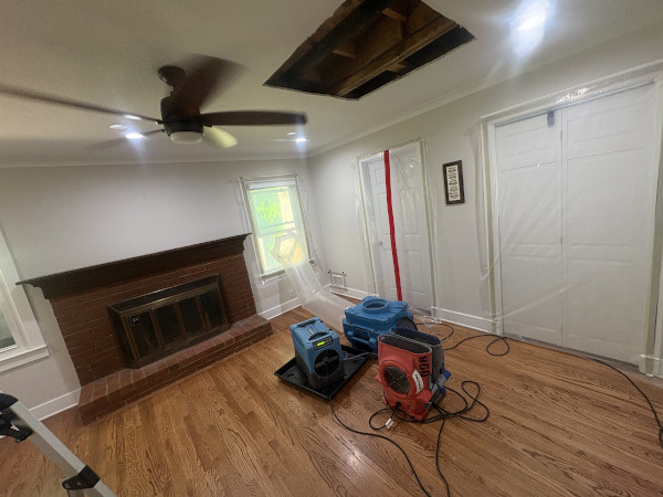 Living room at home on Weatherstone Pkwy, Marietta GA with brick fireplace, ceiling fan, blue and red air movers on hardwood, plastic containment over doors and windows, and large ceiling hole exposing damaged joists. Relief Remediation drying chamber after ceiling demolition.